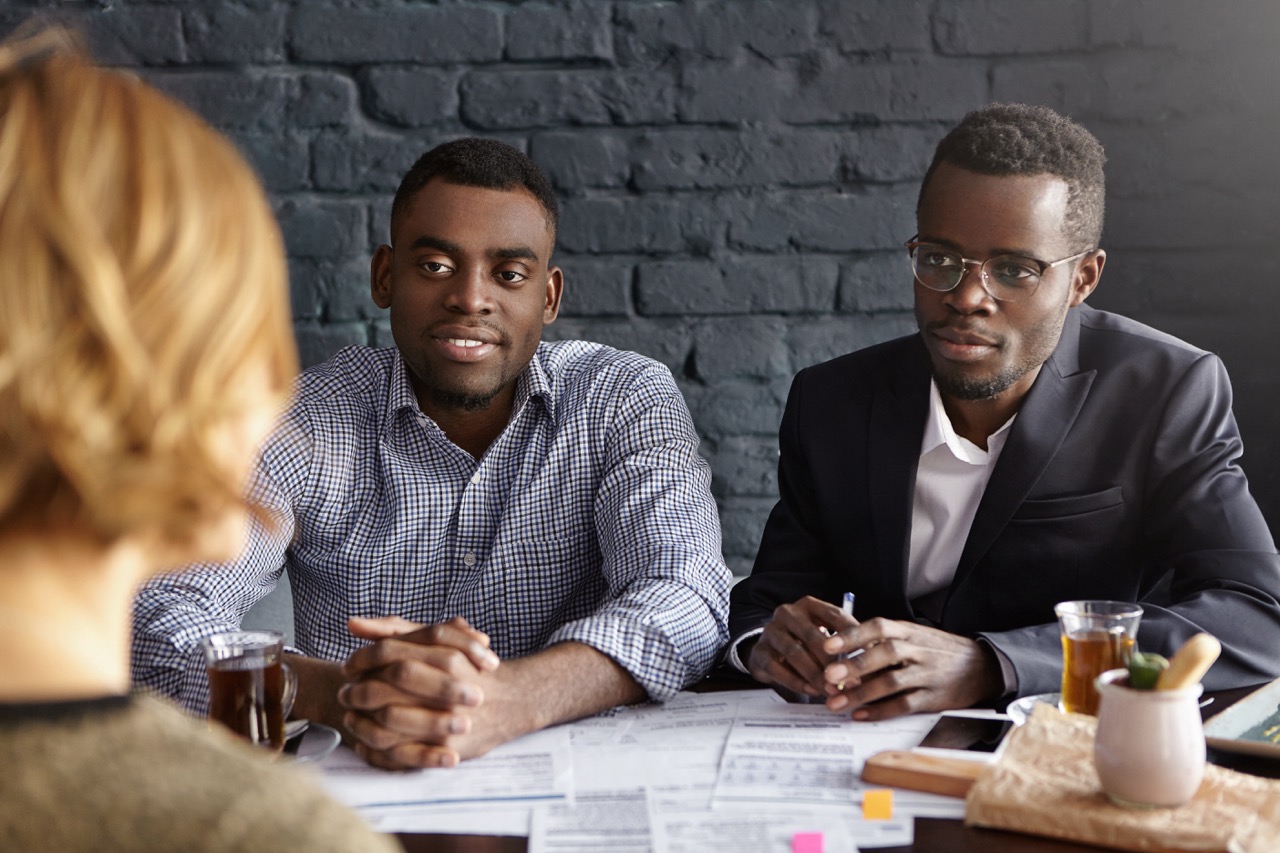 Human resources, career, recruitment and business. Two confident and successful African HR managers interrogating young female candidate during job interview, sitting in office with papers on desk